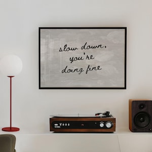 May include: A framed artwork with the text "slow down, you're doing fine" hangs above a record player and speaker. A red floor lamp with a white globe shade stands to the left. The record player is brown with silver knobs.