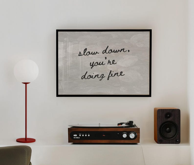 May include: A framed artwork with the text "slow down, you're doing fine" hangs above a record player and speaker. A red floor lamp with a white globe shade stands to the left. The record player is brown with silver knobs.