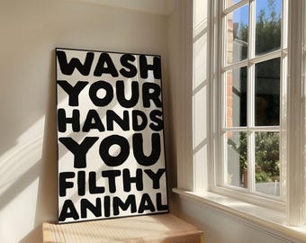 May include: A rectangular wall art print in a thin black frame featuring bold, black, hand-lettered text on a white background that reads: WASH YOUR HANDS YOU FILTHY ANIMAL. The typography is thick, rounded, and sans-serif, filling the entire frame.