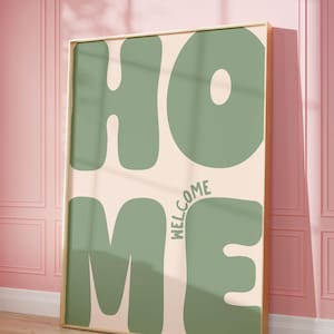 Puede incluir: Una impresión enmarcada con la palabra "HOME" en letras grandes, verdes y con forma de burbuja. Las letras están sobre un fondo rosa claro. La palabra "WELCOME" está escrita en una fuente más pequeña en la esquina inferior derecha de la impresión.