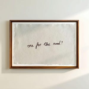 以下が含まれることがあります： 明るい灰色の背景に、手書きのフレーズ「one for the road？」がダークブラウンのインクで書かれた額装されたアートワーク。 アートワークは濃い茶色の木製フレームに収められており、ミニマリストな美学を作り出しています。