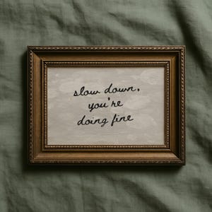 May include: A framed print with the words "slow down, you're doing fine" in black script. The frame is a dark gold colour with ornate detailing. The background of the print is a light grey colour.