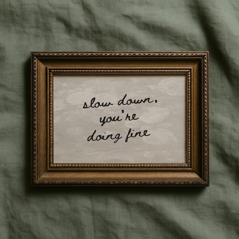 May include: A framed print with the words "slow down, you're doing fine" in black script. The frame is a dark gold colour with ornate detailing. The background of the print is a light grey colour.