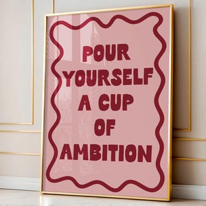 May include: A framed art print with the text "POUR YOURSELF A CUP OF AMBITION" in burgundy letters on a pink background. The print is enclosed in a gold frame with a burgundy wavy border.