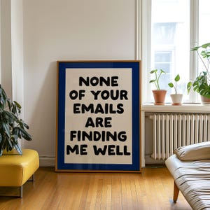 May include: A framed art print with the text "NONE OF YOUR EMAILS ARE FINDING ME WELL" in bold black letters. The artwork has a white background with a blue border and a wooden frame. The print is leaning against a wall.