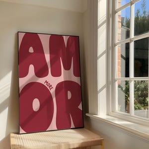 Könnte beinhalten: Ein gerahmter Kunstdruck mit dem Wort "AMOR" in großen, fetten, burgunderfarbenen Buchstaben auf hellrosa Hintergrund. Das Wort "MORE" ist in kleineren burgunderfarbenen Buchstaben geschrieben. Das Kunstwerk wird auf einer Holzbank in der Nähe eines Fensters ausgestellt.