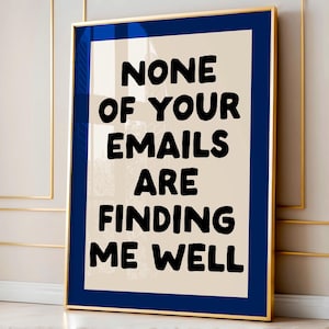 May include: A framed art print with a gold frame and a navy blue mat. The artwork features the text "NONE OF YOUR EMAILS ARE FINDING ME WELL" in bold black letters on a cream-colored background. The print is displayed against a neutral wall.