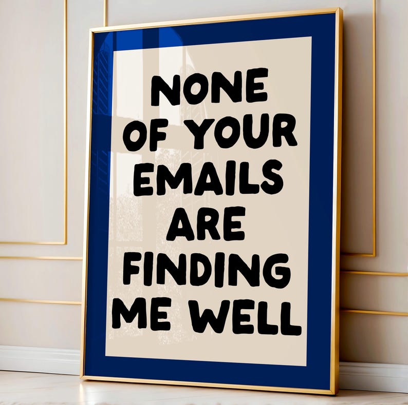 May include: A framed art print with a gold frame and a navy blue mat. The artwork features the text "NONE OF YOUR EMAILS ARE FINDING ME WELL" in bold black letters on a cream-colored background. The print is displayed against a neutral wall.