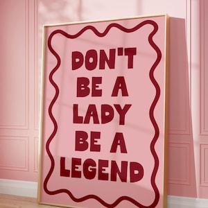 May include: A framed pink poster with the words "DON'T BE A LADY BE A LEGEND" in burgundy lettering. The poster has a gold frame and is leaning against a pink wall and wooden floor.