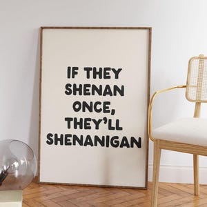 May include: A framed print with the text "IF THEY SHENAN ONCE, THEY'LL SHENANIGAN" in bold black letters. The print is in a wooden frame and is leaning against a white wall. A chair with a gold frame and a glass vase are also in the image.