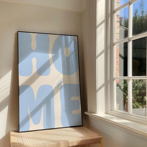 Pode incluir: Uma impressão de parede retangular emoldurada com fundo creme e letras grandes, arredondadas e azul-claras em estilo bolha que formam a palavra HOME. O texto menor, azul e sem serifa SWEET está posicionado entre as letras O e M.