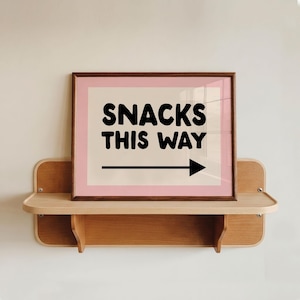Op de afbeelding: Een rechthoekige ingelijste print met een lichtroze rand en een crèmekleurig midden. De tekst SNACKS THIS WAY is gedrukt in dikke zwarte letters, vergezeld van een zwarte horizontale pijl die naar de rechterkant van het frame wijst.