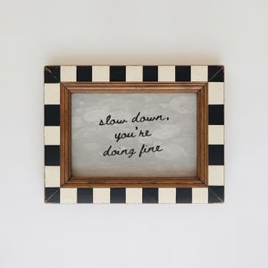 May include: A rectangular wooden picture frame with a black and white chequered border. The frame contains a light grey background with the handwritten text "slow down, you're doing fine."