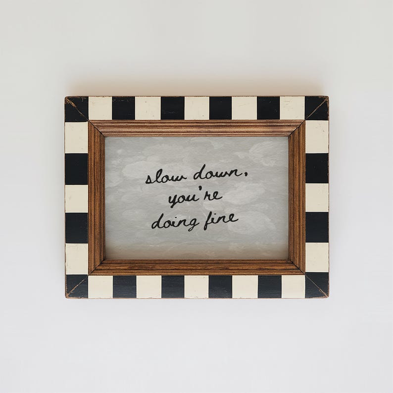 May include: A rectangular wooden picture frame with a black and white chequered border. The frame contains a light grey background with the handwritten text "slow down, you're doing fine."