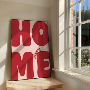 May include: A framed art print featuring the word "HOME" in large, bold, red letters. The word "WELCOME" is printed vertically in smaller red letters. The print is set against a light pink backdrop.