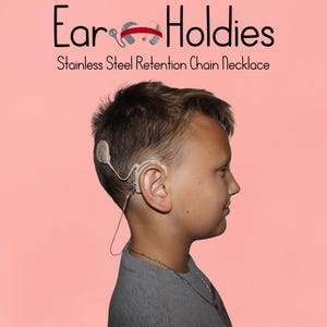 May include: A young person wearing a hearing aid with a stainless steel retention chain necklace. The image has the text "Ear Holdies" and "Stainless Steel Retention Chain Necklace" on a pink background.