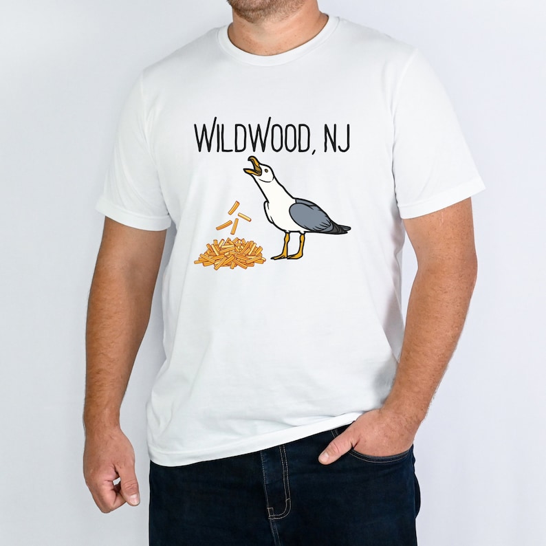 Op de afbeelding: Wit T-shirt met een afbeelding van een meeuw die frietjes eet en de tekst "WILDWOOD, NJ".