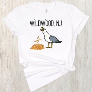 Op de afbeelding: Wit T-shirt met zwarte tekst die "Wildwood, NJ" zegt en een cartoon zeemeeuw met een stapel friet.