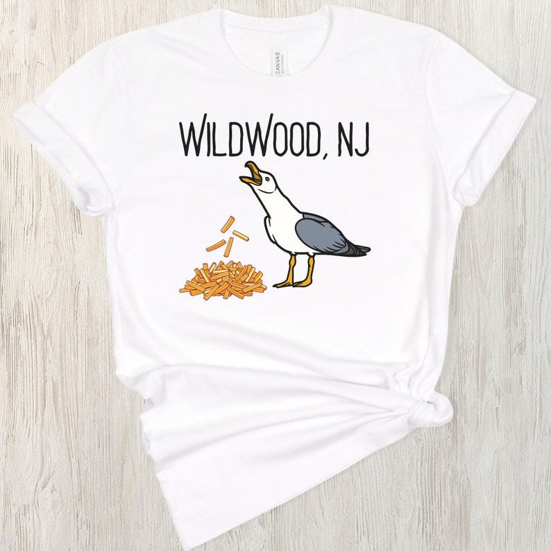 Op de afbeelding: Wit T-shirt met zwarte tekst die "Wildwood, NJ" zegt en een cartoon zeemeeuw met een stapel friet.
