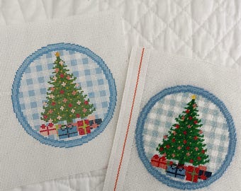 May include: Two needlepoint canvases with blue and white gingham backgrounds. Each canvas features a circular design with a green Christmas tree and presents.