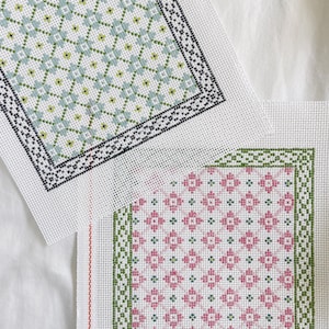 May include: Two white canvas needlepoint projects with different patterns. The top project has a blue and green geometric pattern with a black border. The bottom project has a pink and green geometric pattern with a green border.