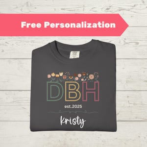 May include: Dark gray t-shirt with floral design above the letters "DBH" in green, yellow, and pink. The text "est. 2025" and "Kristy" are below the letters. A red banner at the top says "Free Personalization".