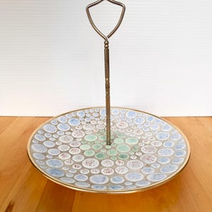 May include: A single-tier serving tray featuring a round ceramic plate with a decorative pattern of small, multicolored circles in shades of blue, green, and reddish-brown. The plate has a gold-toned metal rim and a central gold-toned metal handle.