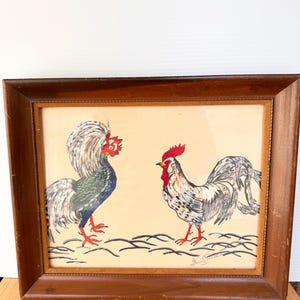 May include: A framed painting featuring two roosters facing each other. The left rooster has white, blue, and green plumage with a red comb. The right rooster is white with black and grey markings and a red comb. A signature is visible at the bottom.