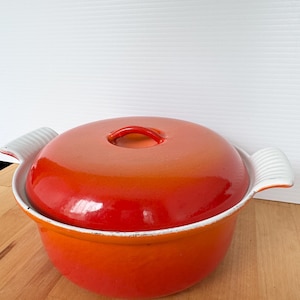 May include: An orange enameled cast iron Dutch oven with a matching lid. The pot has white handles and a white rim. The lid has a small loop handle. The pot sits on a wooden surface.