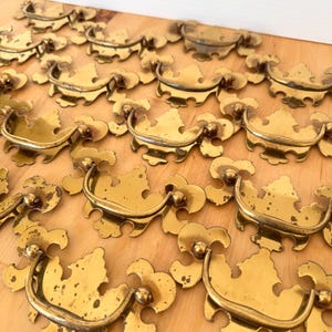 May include: A collection of vintage brass drawer pulls arranged on a light wood surface. Each pull features a decorative, ornate design with a curved handle and two small ball accents. The brass has a slightly aged patina.