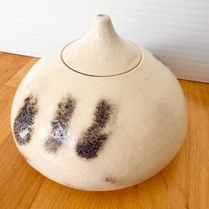 May include: Cream-colored ceramic jar with a lid, featuring dark speckled markings. The jar has a rounded body and a conical top. The surface has a crackled texture. The jar is sitting on a wooden surface.