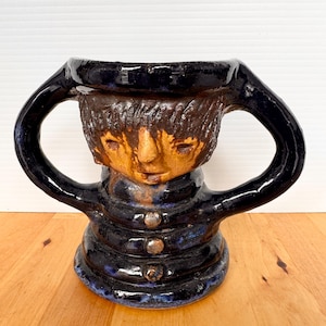 May include: A ceramic mug with a face design. The mug features a dark blue rim and handles, with a brown face and hair. The body of the mug is dark blue with three button details. The mug is sitting on a wooden surface.