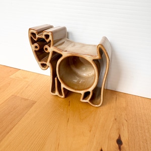 May include: A ceramic cow-shaped planter in a light brown color. The planter has a large circular opening in the center and is designed with cut-out details to resemble a cow. The planter is sitting on a wooden surface.