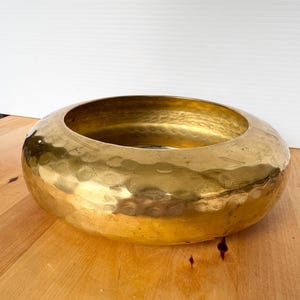 May include: A round, low-profile bowl crafted from polished brass with a distinct hammered texture across its exterior surface. The vessel features a wide, curved rim that slopes inward toward a smooth, circular opening at the center.