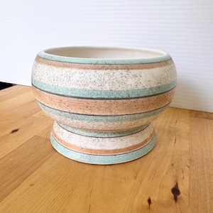 May include: A ceramic bowl with a pedestal base featuring horizontal stripes in shades of light green and tan. The surface has a speckled texture throughout, and the interior of the bowl is a solid off-white color with a smooth, rounded rim.