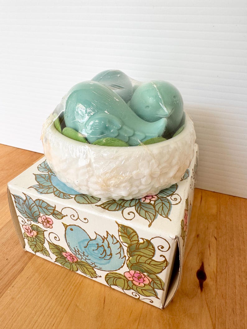 Puede incluir: Un juego de tres jabones de p&aacute;jaros azules anidados en un nido blanco texturizado, envueltos en pl&aacute;stico transparente. El nido se encuentra sobre una caja decorativa con un dise&ntilde;o de p&aacute;jaro azul y floral.