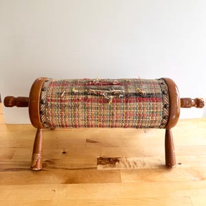 May include: A cylindrical footstool featuring a woven fabric cover with a multicolored striped pattern, secured by metal studs to two polished wooden end caps. The structure is supported by four turned wooden legs with visible wear on the finish.
