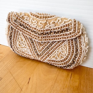 May include: A vintage beaded clutch in cream and gold tones. The rectangular bag features intricate beadwork in a floral pattern. The flap closure is also beaded. The clutch is resting on a wooden surface.