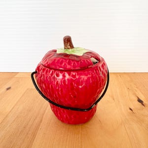 May include: A ceramic strawberry-shaped container with a lid and a black handle. The container is a vibrant red with green leaves and a brown stem on top. The container sits on a light-colored wooden surface.