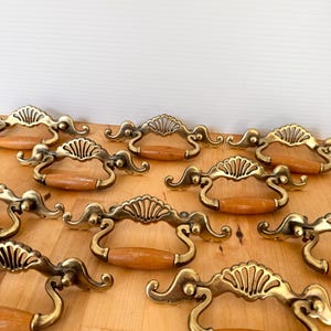 May include: A collection of vintage brass drawer pulls with wooden handles. The brass pulls have a decorative fan-shaped design and are arranged on a light-colored wooden surface. The handles are approximately 4 inches long.