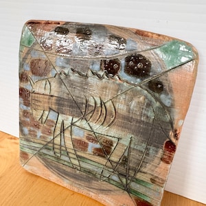 May include: A handcrafted ceramic wall art piece with a unique, abstract design. The artwork features a textured surface with a fish-like figure and geometric lines. Earthy tones of brown, green, and blue are visible.
