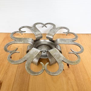May include: A circular metal candle holder with a brushed silver finish. The design features eight curved, petal-like arms extending outward from a central, recessed circular base. The metal has a matte, slightly weathered texture throughout.
