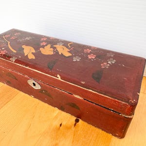 May include: A rectangular, antique wooden box with a dark red finish. The box is adorned with floral designs in gold, pink, and silver. It features a metal keyhole and shows signs of wear, suggesting its age and history. The box is likely used for storage.