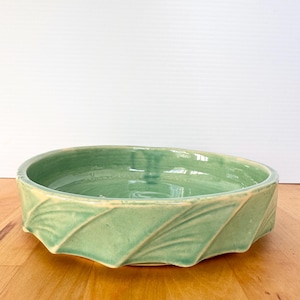 May include: A round, shallow ceramic bowl with a glossy light green glaze. The exterior features a series of raised, diagonal ridges that create a pleated or leaf-like pattern around the circumference of the vessel.