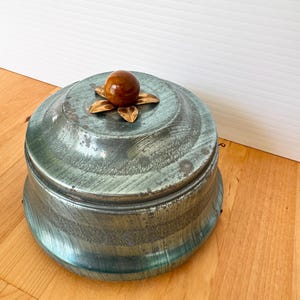 May include: A round, antique-style metal box with a teal and silver finish. The lid is adorned with a brown, spherical knob and gold-colored petal accents. The box sits on a wooden surface.