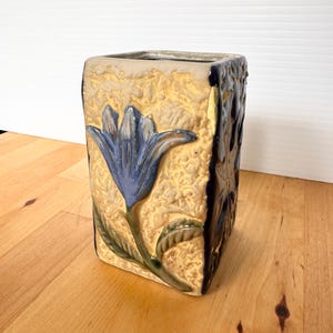 May include: A rectangular ceramic vase with a textured beige surface featuring a raised blue flower design on the front. The side of the vase is finished in a dark, glossy blue glaze. The top opening is square, and the piece has a sturdy base.