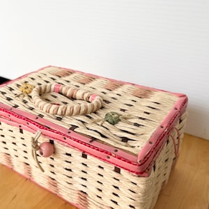 May include: A pink and white woven wicker box with a pink ribbon handle and a pink button closure. The box has a light brown wooden base.