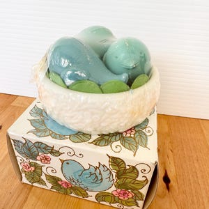 Puede incluir: Un juego de regalo decorativo con tem&aacute;tica de p&aacute;jaros. El juego incluye tres art&iacute;culos azules con forma de p&aacute;jaro en un recipiente blanco con forma de nido con detalles de hojas verdes, encima de una caja con un dise&ntilde;o de p&aacute;jaro y flores.