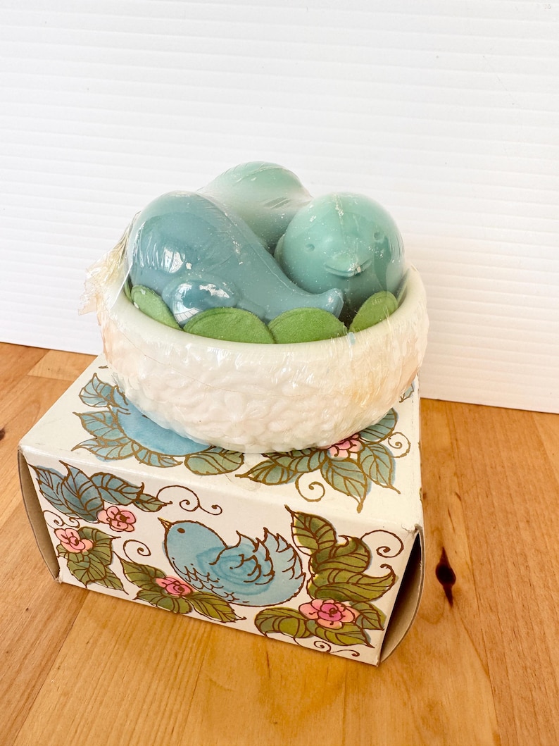 Puede incluir: Un juego de regalo decorativo con tem&aacute;tica de p&aacute;jaros. El juego incluye tres art&iacute;culos azules con forma de p&aacute;jaro en un recipiente blanco con forma de nido con detalles de hojas verdes, encima de una caja con un dise&ntilde;o de p&aacute;jaro y flores.