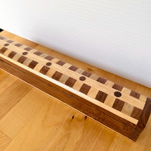 May include: A rectangular wooden box with a decorative inlaid design. The box features alternating bands of light and dark wood, with a repeating pattern of squares and circles. The box is made of various wood tones and sits on a wooden surface.